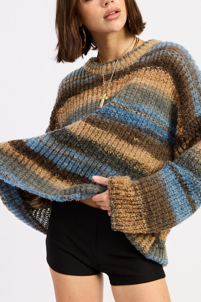 A woman wearing a chunky knit striped sweater with horizontal bands of blue, brown, beige, and gray, paired with black shorts against a light gray studio background.