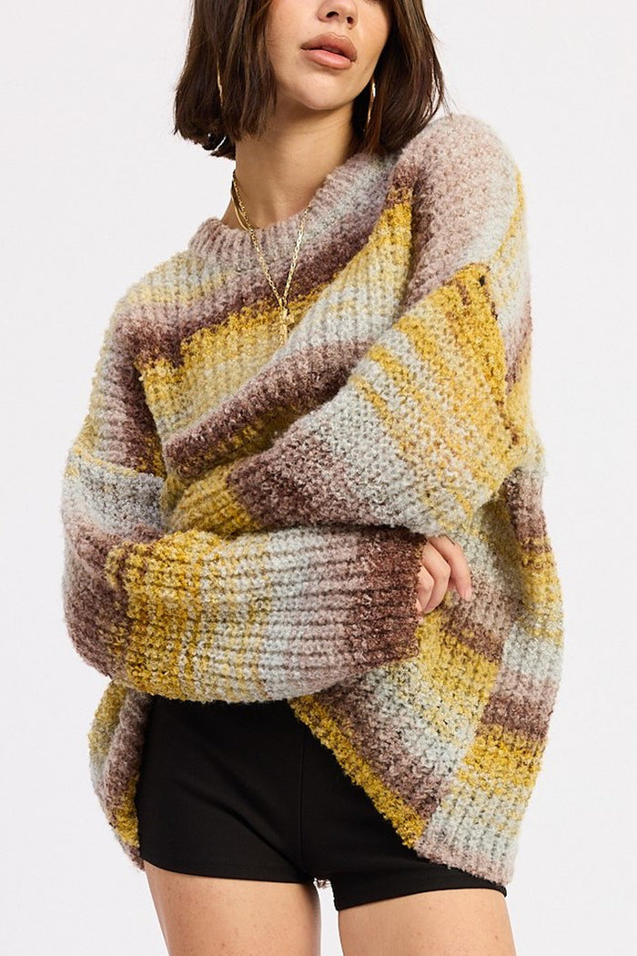 A woman wearing a chunky knit sweater with horizontal stripes in mustard yellow, brown, beige, and light gray, paired with black shorts against a plain white background.