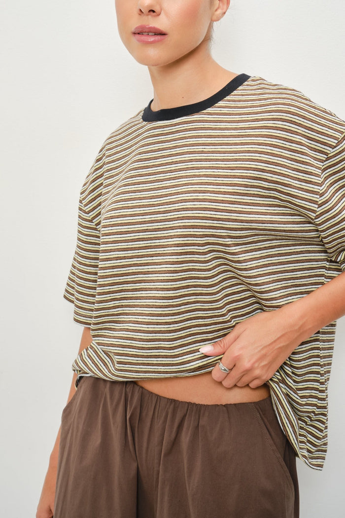 A close-up of a person wearing a loose short-sleeve striped top with a black crew neck, paired with brown pants, against a plain light background.
