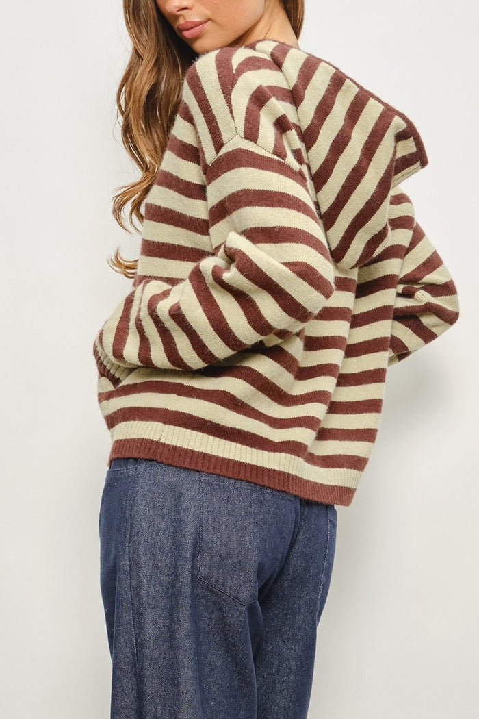 A woman wearing a chunky knit sweater with horizontal brown and cream stripes, paired with blue jeans, shown from a side-back angle against a plain light background.