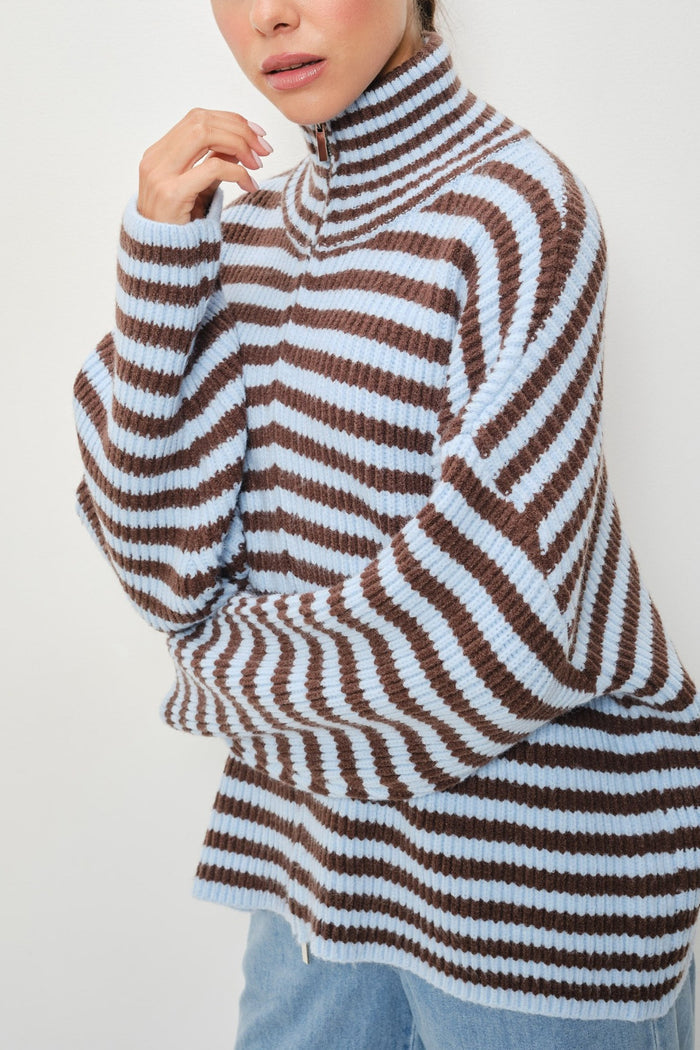 A close-up of a person wearing a ribbed knit mock-neck sweater with horizontal brown and light blue stripes, paired with light blue jeans against a plain light background.
