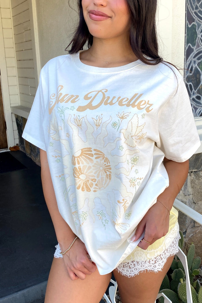 Woman wearing a white t-shirt with gold text and patterns, standing outdoors.