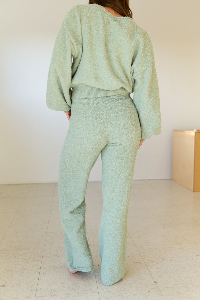 Person wearing a light green outfit standing in a minimalistic room with a white wall and wooden box.