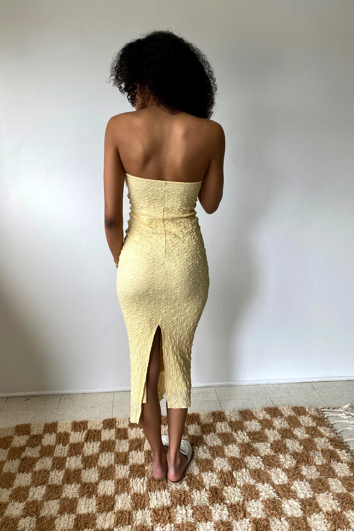 Woman wearing a strapless yellow dress with a slit, standing on a checkered rug.